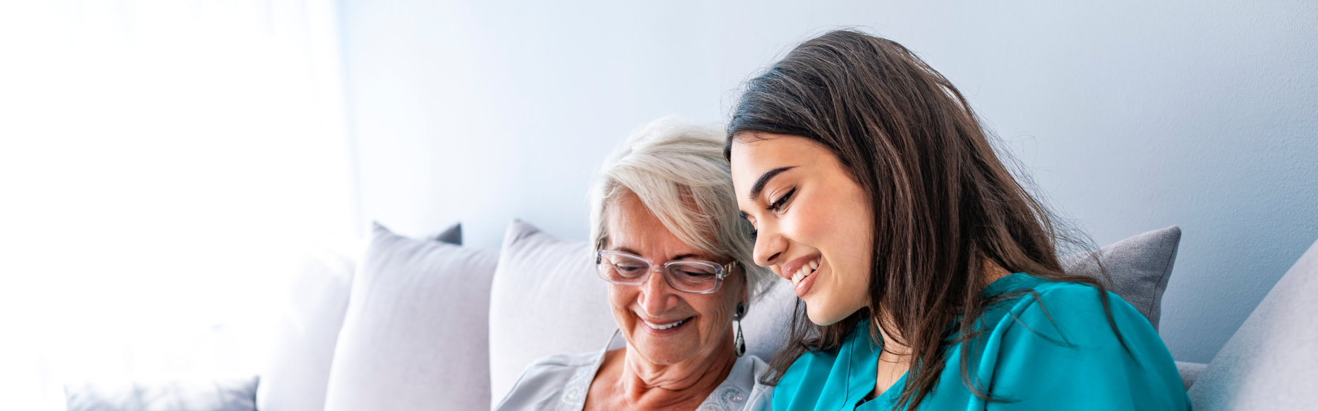 Caregiver and elderly woman looking at something while sitting on the couch